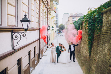 Newlyweds and their friends raise their hands with colors smoke. Running with smoke bombs. Red and blue colors. Wedding couple with color smoke.