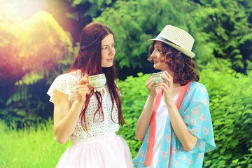 Obraz premium Two brunette girls drink tea and talk in the park on a picnic. Two friends communicate at nature. Girlfriends in the forest. Toned photo of two models in the park