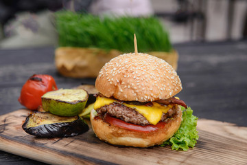 Cheeseburger on the wooden table outdoors. served with grilled vegetables. Home made fast food from fresh, tasty ingredients. Hamburger with crispy beckon and tomato inside