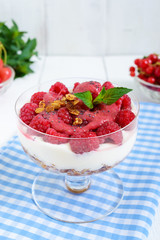 Sweet creamy dessert with granola, cream cheese, fresh raspberries in a glass bowl on a white wooden background.