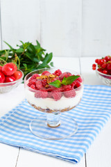 Sweet creamy dessert with granola, cream cheese, fresh raspberries in a glass bowl on a white wooden background.