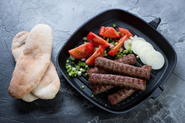 Cast-iron grill pan with balkan roasted cevapi sausages, fresh tomatoes, onion and pita bread, horizontal shot