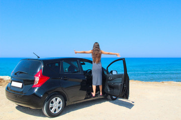 A woman with her arms up next to a rented car with a view of the sea