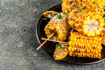 Summer food. Ideas for barbecue and grill parties. Grilled corn grilled on fire. With a sprinkle of cheese (mexican elotes), hot chili pepper, lemon. On a dark stone table, black plate Copy space