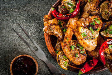 Summer food. Ideas for barbecue, grill party. Chicken legs, wings grilled, fried on fire. With hot chili pepper, lemon and bbq sauce. Dark stone table, on black plate Copy space