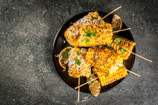 Summer food. Ideas for barbecue and grill parties. Grilled corn grilled on fire. With a sprinkle of cheese (elotes), hot chili pepper, lemon. On a dark stone table, black plate Copy space top view