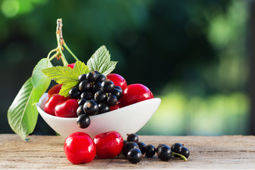 Fresh black currant and red cherry on a branch with leaves in a porcelain saucer. Handful of berries on the wooden table and blurred background. Shallow depth of field