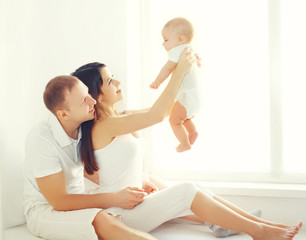 Happy family, mother and father playing with baby home in white room near window