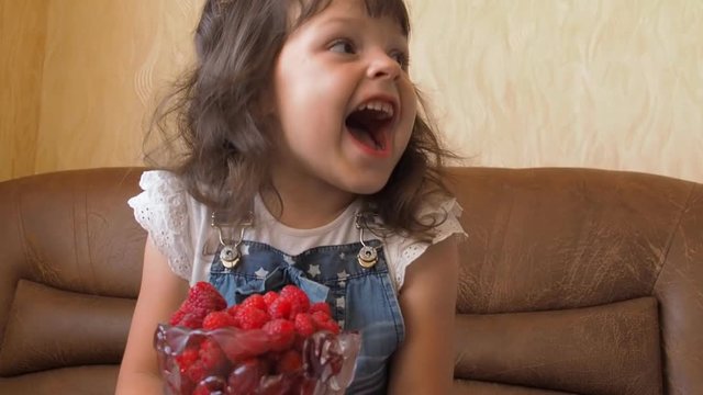 Little girl looks at a raspberry. The child eats raspberries.