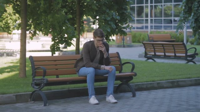 Young attractive man in glasses, in park on bench, smoking a cigarette