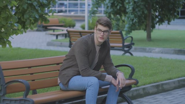 Young attractive man in glasses, in park on bench, smoking a cigarette