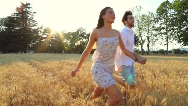 Beautiful Young Couple running and having fun  in a wheat field / A man and woman in love in the middle of nature holding hands smiling in sunset light surrounded by trees / slow-mo / 720p / steady-ca