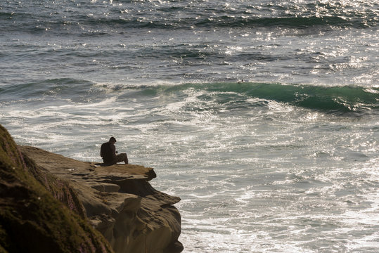 Man Sitting On Cliff Overlooking The Pacific Ocean Looking At His Cellphone 