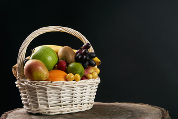 Assortment of fresh raw fruits in wicker basket on wooden stump isolated on black background. Healthy eating concept.