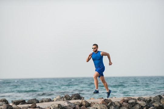 Strong sportsman in blue shorts and shirt jogging along coast