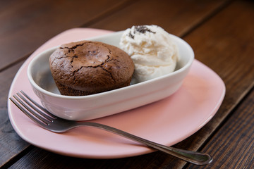 Chocolate lava cake with ice cream covered with poppy seeds, in a white bowl, on a pink plate, fork near by. Wooden table. Selective focus, small depth of field. 