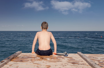 Fototapeta na wymiar Boy with diving mask, sitting on a wooden jetty at sea beach. Daydreaming concept.