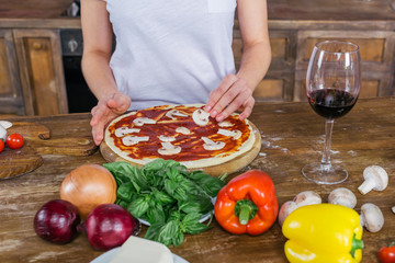 Cropped shot of young woman putting mushrooms on raw pizza while standing at kitchen table