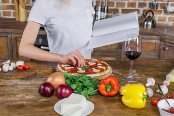Cropped shot of young woman holding blank cookbook while preparing pizza at kitchen