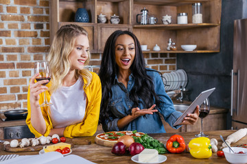 Cheerful multiethnic women using digital tablet while cooking together in kitchen