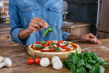 Cropped shot of woman putting basil leaves on raw pizza