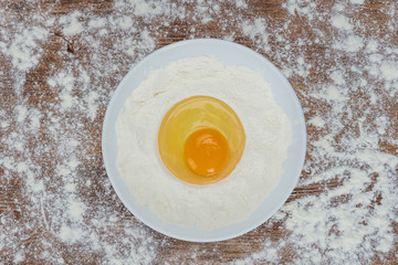 Top view of raw broken egg with flour on plate on wooden table top