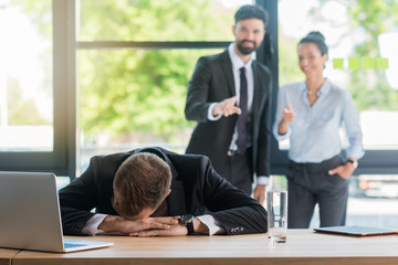 tired businessman at sitting workplace with laptop in office while colleagues standing behind