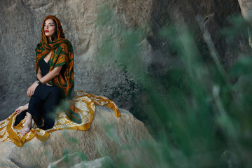 Obraz na płótnie Canvas Beautiful Muslim woman with oriental scarf on head sitting on cliff on a mountain background