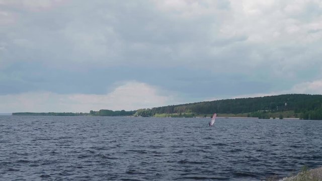Caucasian man in a black hydro-suit riding windsurf with a red sail against the backdrop of a forest shore in Tolyatti, Russia