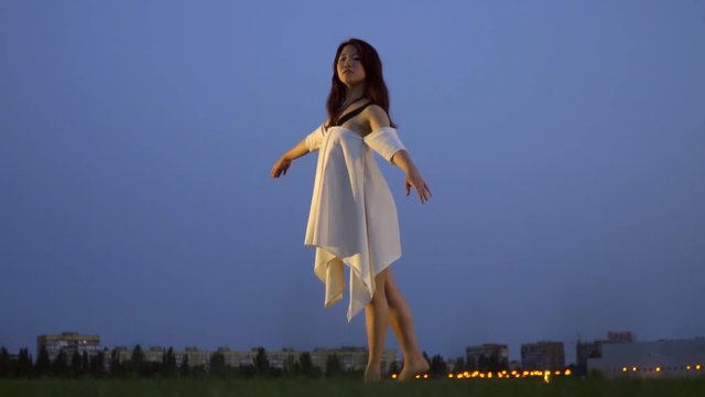 Flexible ballerina in white dress posing stretching her leg against the background of the evening sky, slow motion