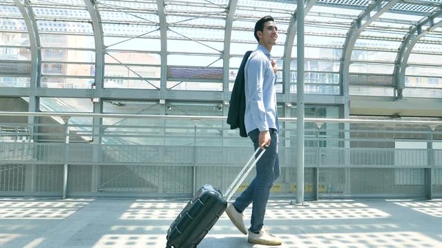Young handsome businessman (student) in a blue shirt, comes with luggage, holds a jacket, at the station, at airport. Concept: new business, traveling the world, communication, contacts, hand luggage.