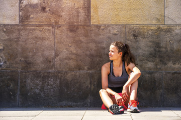 Shot of fit runner siting against a wall outdoors. Sporty woman relaxing after running exercise.