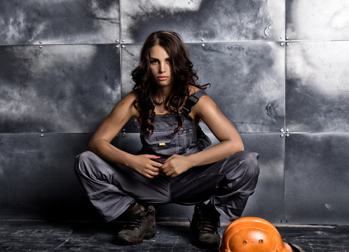 Beautiful sexy worker with a pickaxe and helmet. Seductive and beautiful woman miner sitting on a floor on steel background