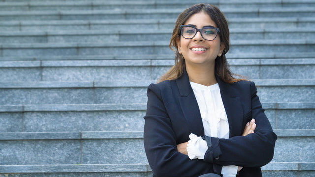 Portrait of young beautiful business woman (student) in suit, glasses, smiling, happy, walking down stairs, steps, on building background. Concept: new business, communication, Arab, banker, manager.