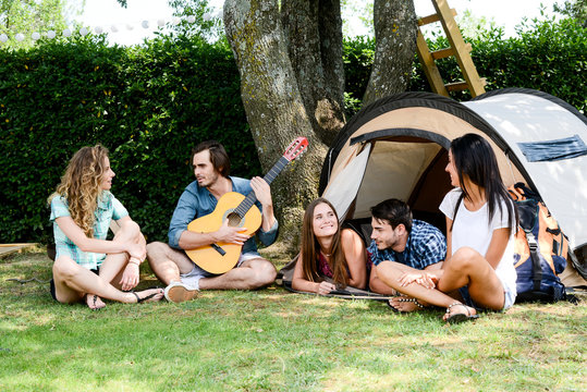 group of five young people playing guitar in a camp site during summer holiday vacation adventure trip