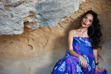 Beauty Fashion Portrait. Beautiful Woman with Curly Hair with colorful elegant long dress Sits in a cave