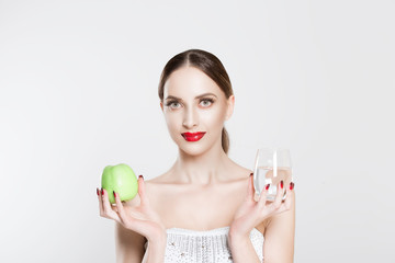 Attractive young woman looking at camera, holding in her hands green apple and glass of water promoting healthy lifestyle, diet and sport, isolated on white background. Face expression, body language.