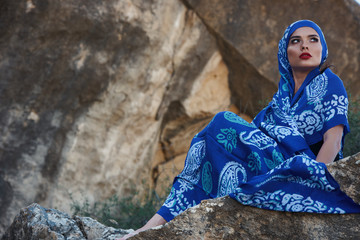 Gorgeous young long hair brunette beauty with slanted head wearing oriental colorful scarf Sits on rocks on a mountain background