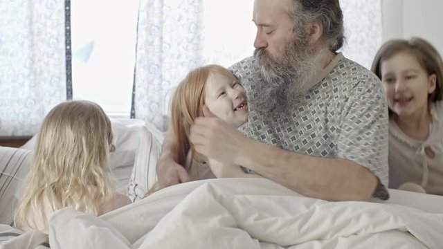 PAN with slow motion of happy little girls with long hair sitting in parents bed and hugging senior father with grey beard as their mother smiling and lying with their little sibling