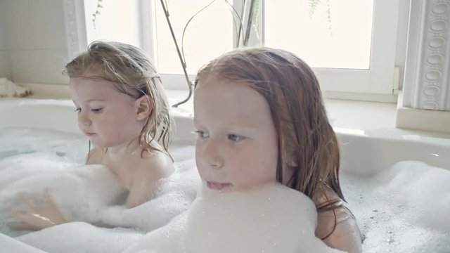 PAN of little girl with wet hair playing with foam in bathtub while her younger brother splashing water and having fun