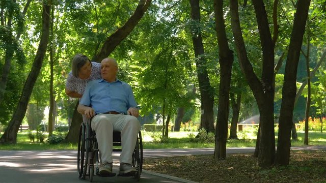 Senior woman pushing husband in wheelchair outside in summer park. 4k footage in slow motion.