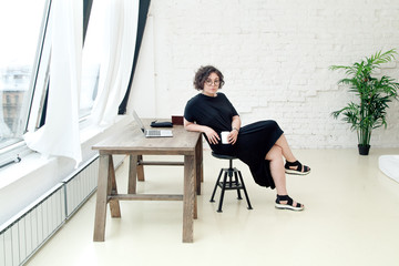 Young plus size female model sitting in front of the table and working on her notebook. Bright modern coworking hall. Composition with copy space.