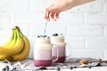 Fototapeta na wymiar Hand Holding a Colorful Straw above Blueberry Coconut Layered Smoothie on the White Brick Background