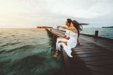 Woman in white long dress is showing to her friend something on horizon during beautiful sunset on Maldives island while they both sitting on wooden bridge and smiling, ocean villas in background