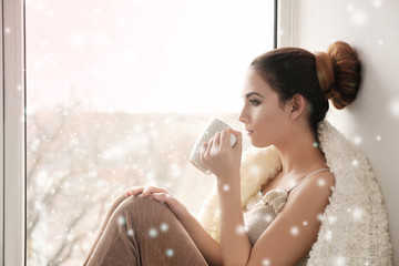 Beautiful young woman drinking coffee while sitting on window sill at home