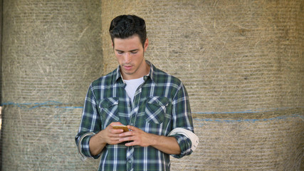 Obraz premium A handsome young farmer (student) is sitting on a haystack, talking on the phone, writing a message, selfie, in a shirt, happy smiling. Concept: communication, ecology, agriculture, hay, contacts.