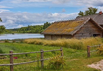 Fototapeta na wymiar Rural landscape/A wooden rural house with a roof covered with moss. In the foreground there is a wooden fence made of poles. In the background a pond. Russia, Pskov region, nature, landscape
