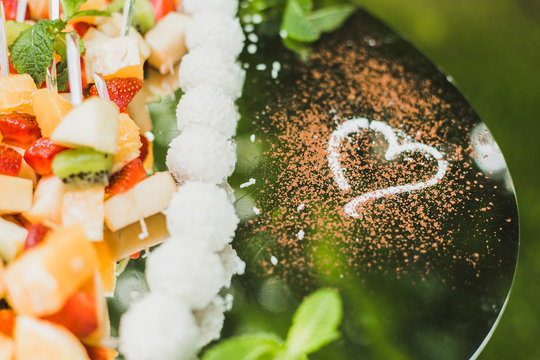 Catering for romantic party. Closeup of various kinds of fruit with cheese and white round chocolate sweets on big tray decorated with heart shape of white color. Horizontal image.