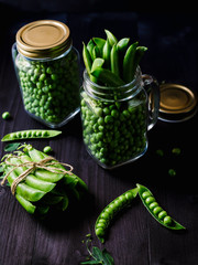 Two full glass jars with fresh mature green peas near bunch of pods tied with rope on black wooden background. Bio healthy food. Green peas, pods, pea leaves and jar on wooden table