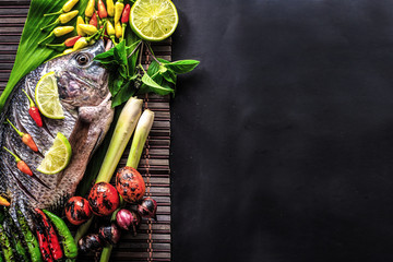 Top view delicious fresh fish and herbs and spices, lemon, tomato, chili and various vegetables on dark wood background, space for texture. Healthy food, diet or cooking concept.